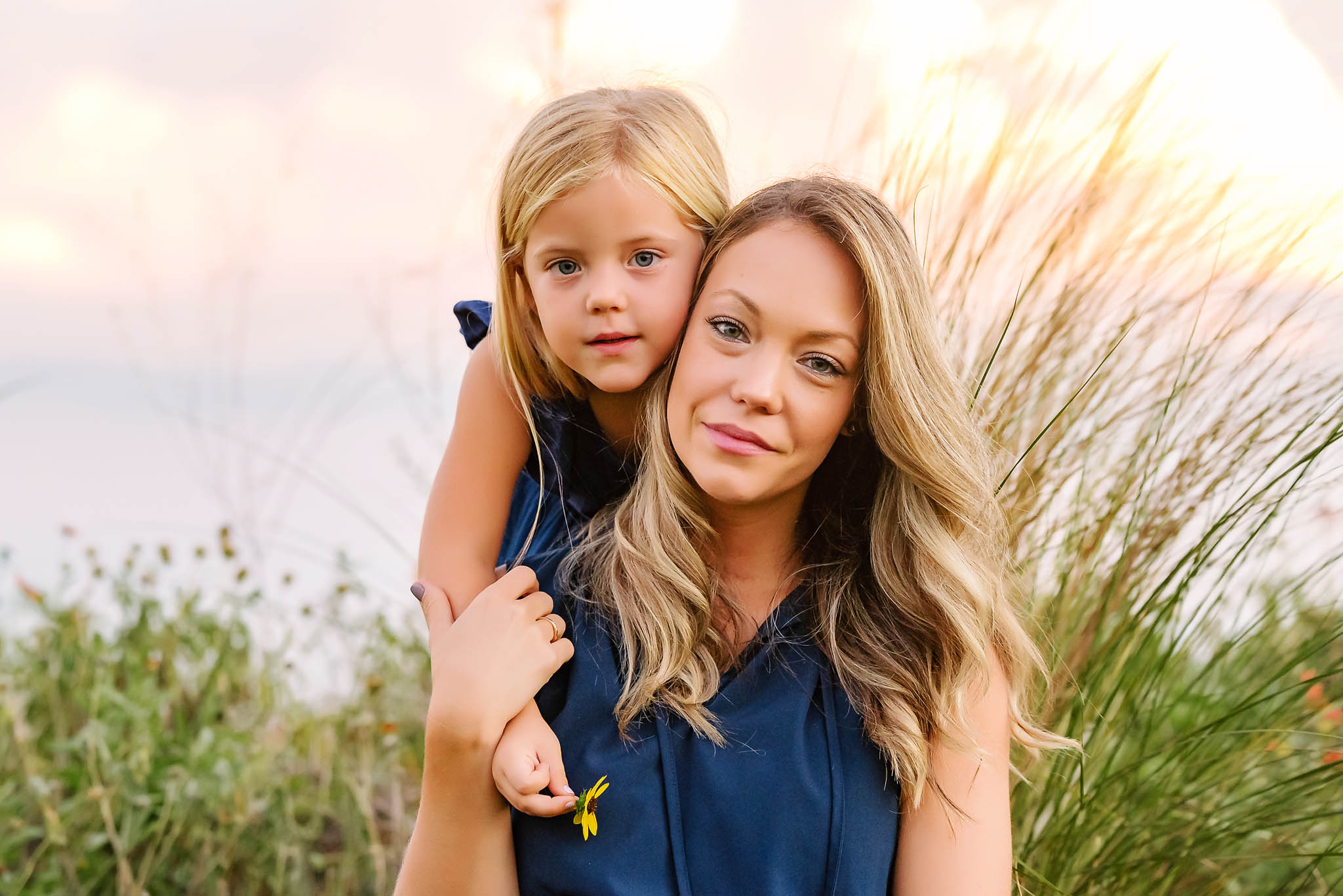 Gorgeous mom and daughter look into camera softly wearing blue and holding flowers