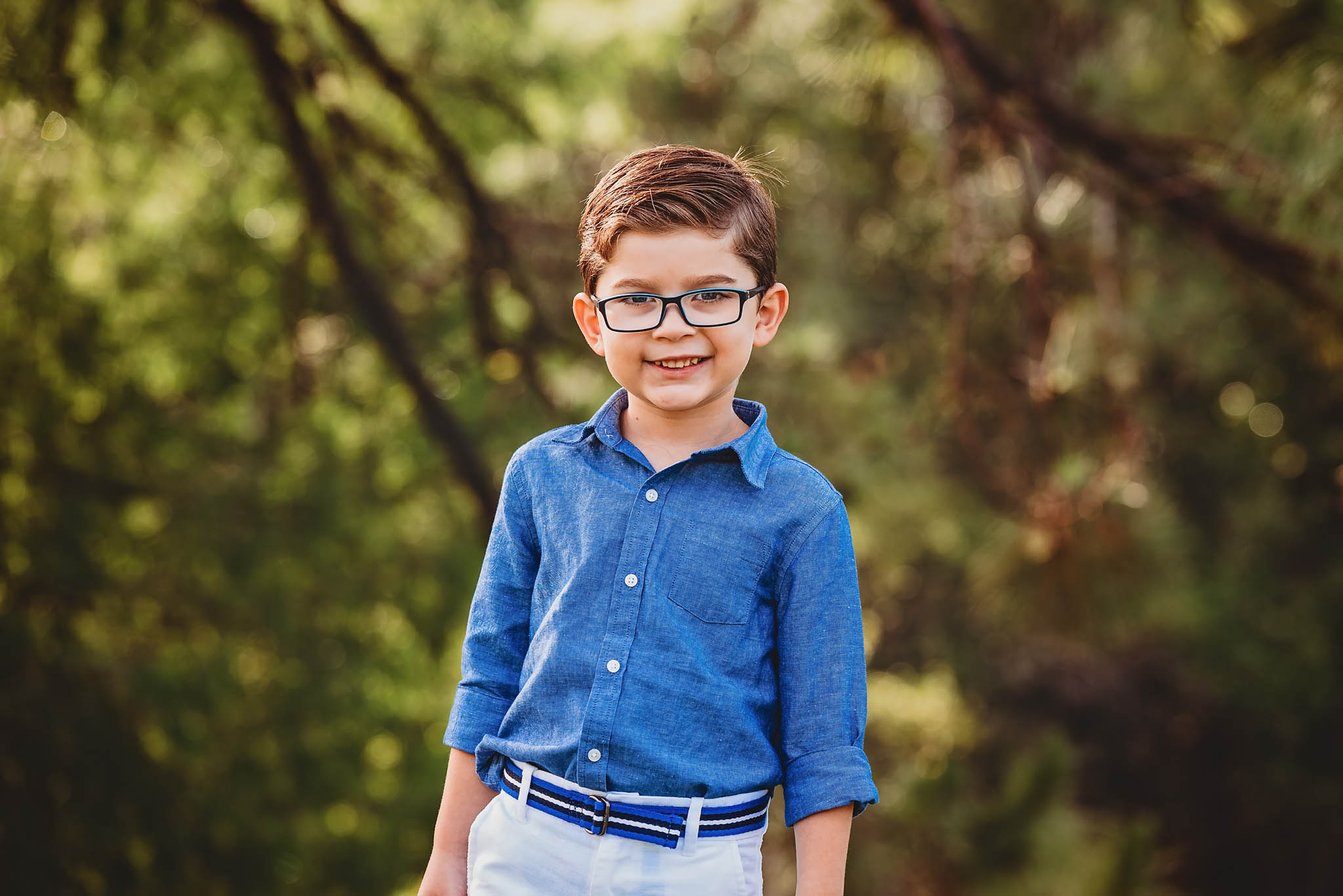 A little boy wearing blue glasses and a blue shirt smiles sweetly into the camera
