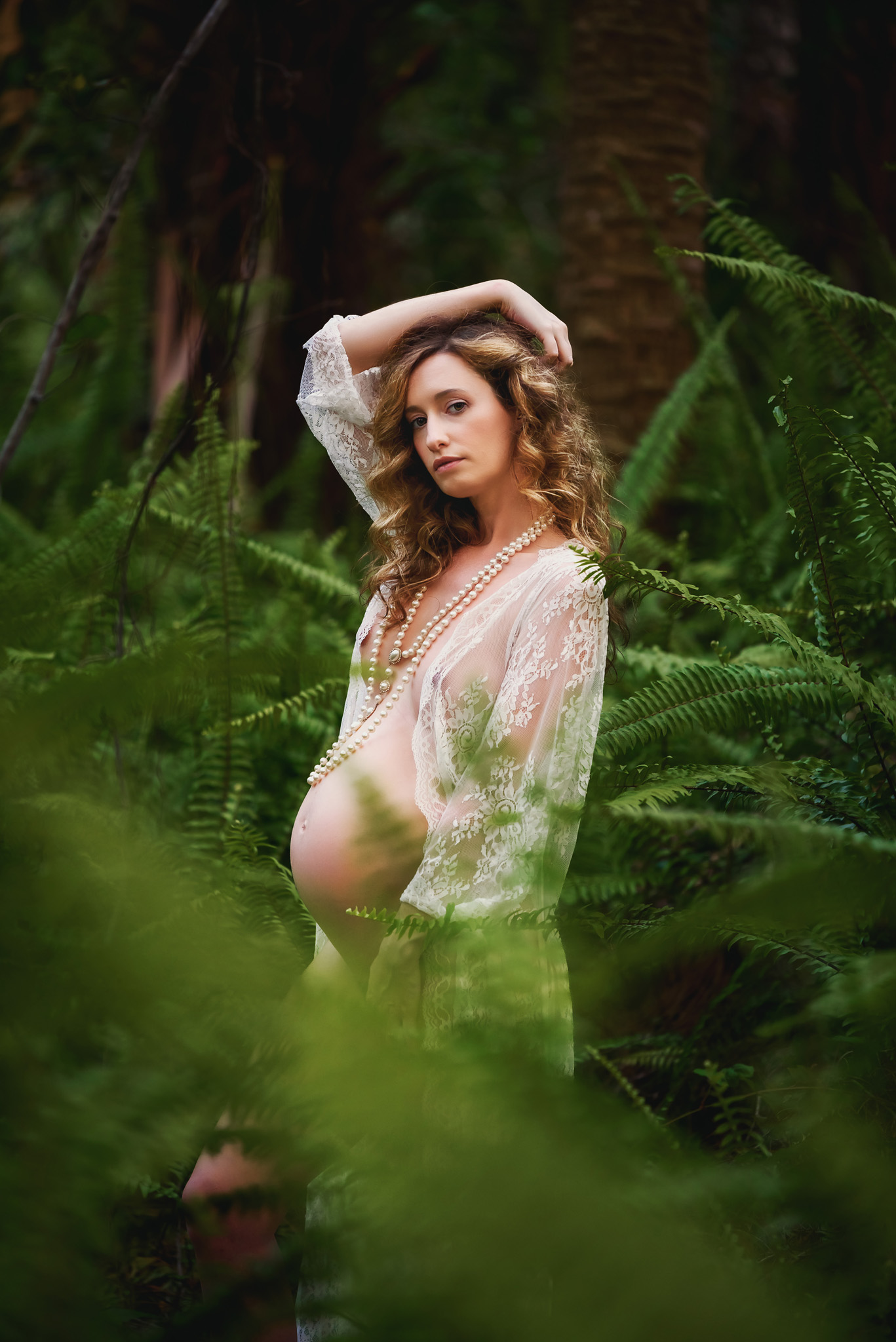 A woman stands in the forest in a white lace robe looking into the camera with an arm over her head during her maternity photoshoot in Fort Myers Florida