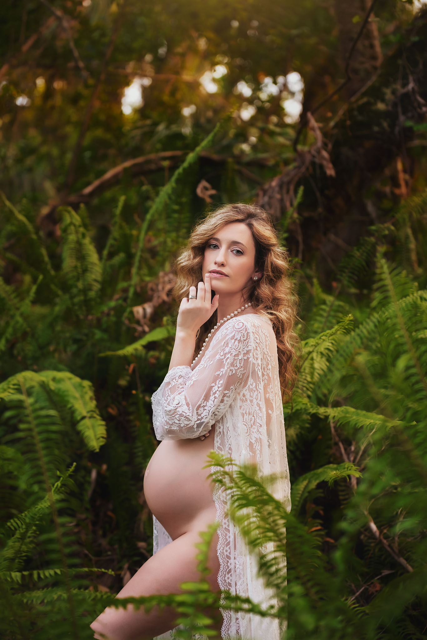 A pregnant woman stands in the ferns in a white lace robe looking into the camera during her maternity session in Fort Myers Florida